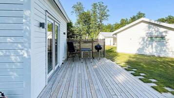 A deck with a grill and chairs, a sliding glass door, and a white building with a sloped roof.