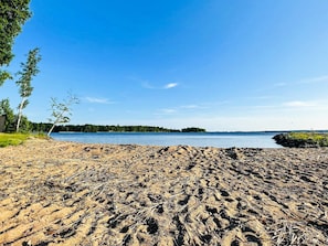 Praia arenosa com vista clara para um lago e uma ilha arborizada ao longe.
