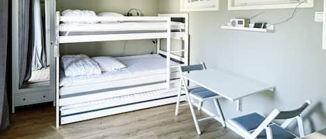 A bunk bed with a desk and chairs, a small shelf with items, and a window with a view of trees.