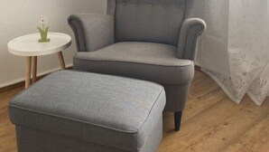 En grå lænestol med tilhørende skammel, et lille rundt bord med en vase, en gulvlampe og et billede i ramme på væggen.