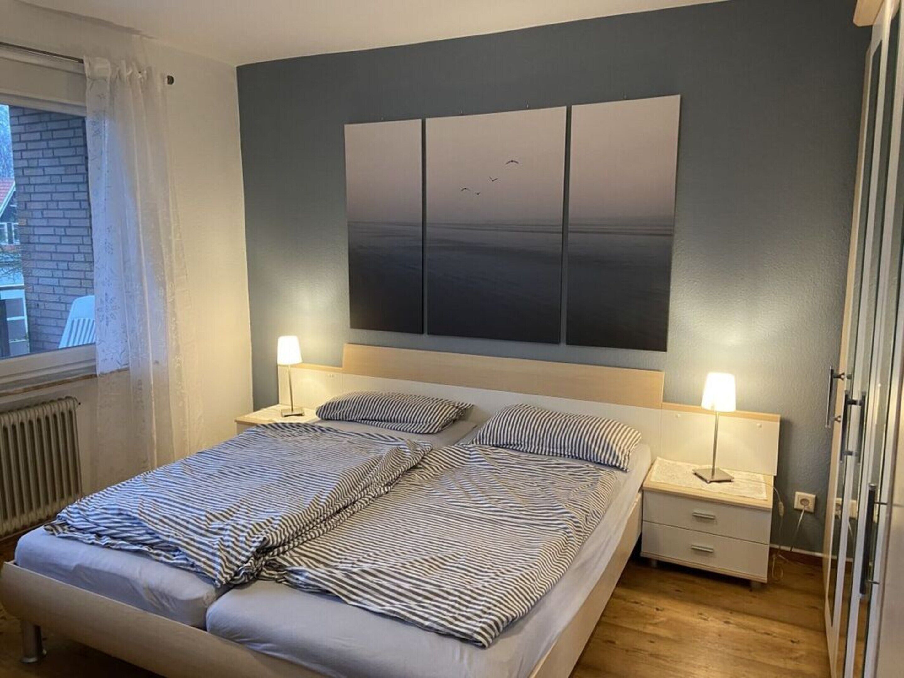 Un dormitorio con cama, dos mesitas de noche con lámparas, un cuadro grande en la pared y una ventana con cortinas.