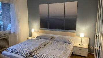 Un dormitorio con cama, dos mesitas de noche con lámparas, un cuadro grande en la pared y una ventana con cortinas.
