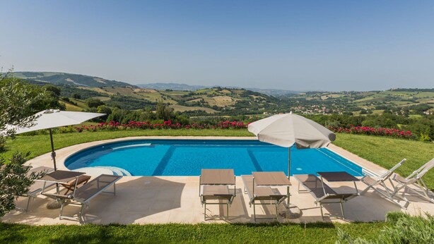 Piscina con sillones de descanso y sombrillas, rodeada de un jardín ajardinado y una vista panorámica de colinas onduladas.