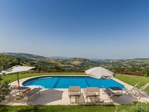 Piscina con sillones de descanso y sombrillas, rodeada de un jardín ajardinado y una vista panorámica de colinas onduladas.
