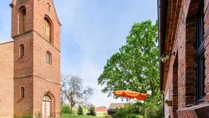 A brick building with a tower, a patio with wooden chairs, and an orange umbrella.