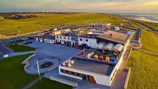 An airport terminal with a modern design, featuring a central building with a curved roof and multiple wings. There are parked cars and a small parking lot in front. - Sun, Mudflats and sea (Wurster Nordseeküste)