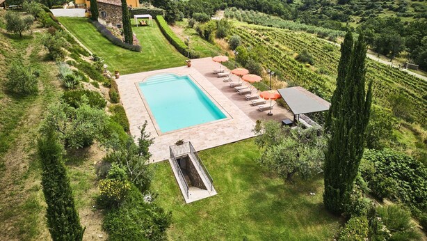 A large rectangular pool with a slide, surrounded by a deck with lounge chairs and umbrellas, set in a lush garden with a view of distant mountains. - Farmhouse in Chianti With Pool and Garden (Cavriglia)