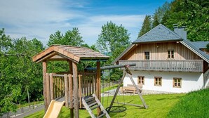 A wooden play structure with a slide and swings, a white house with a balcony, and a grassy yard. - Infanglalm "traunsee" (Altmuenster)