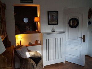 A room with a wicker chair, a white radiator, a door with a round mirror, and a shelf with a lamp and a small decorative item.