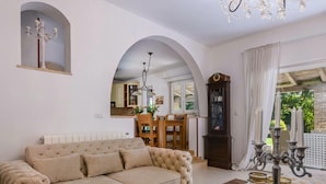 A living room with a beige sofa, wooden coffee table, and a chandelier. There is a dining area with a wooden table and chairs, and a kitchen visible through an archway. - Villa Regenboog (Porec)