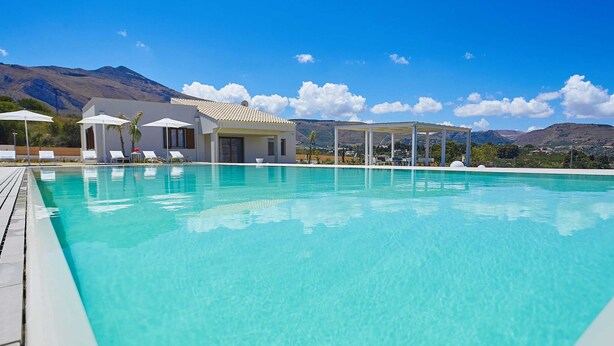 Ein klares, blaues Schwimmbecken mit Liegestühlen und Sonnenschirmen, einem überdachten Sitzbereich und einem Berg im Hintergrund.