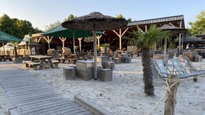 Zona de playa con mobiliario de terraza de madera, sombrillas de paja y un edificio de madera con señalización.