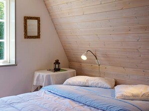 A bedroom with a sloped ceiling, a bed with white and blue bedding, a small bedside table with a lantern, and a wall-mounted lamp. - 4 Person Holiday Home in Hedekas (Hedekas)