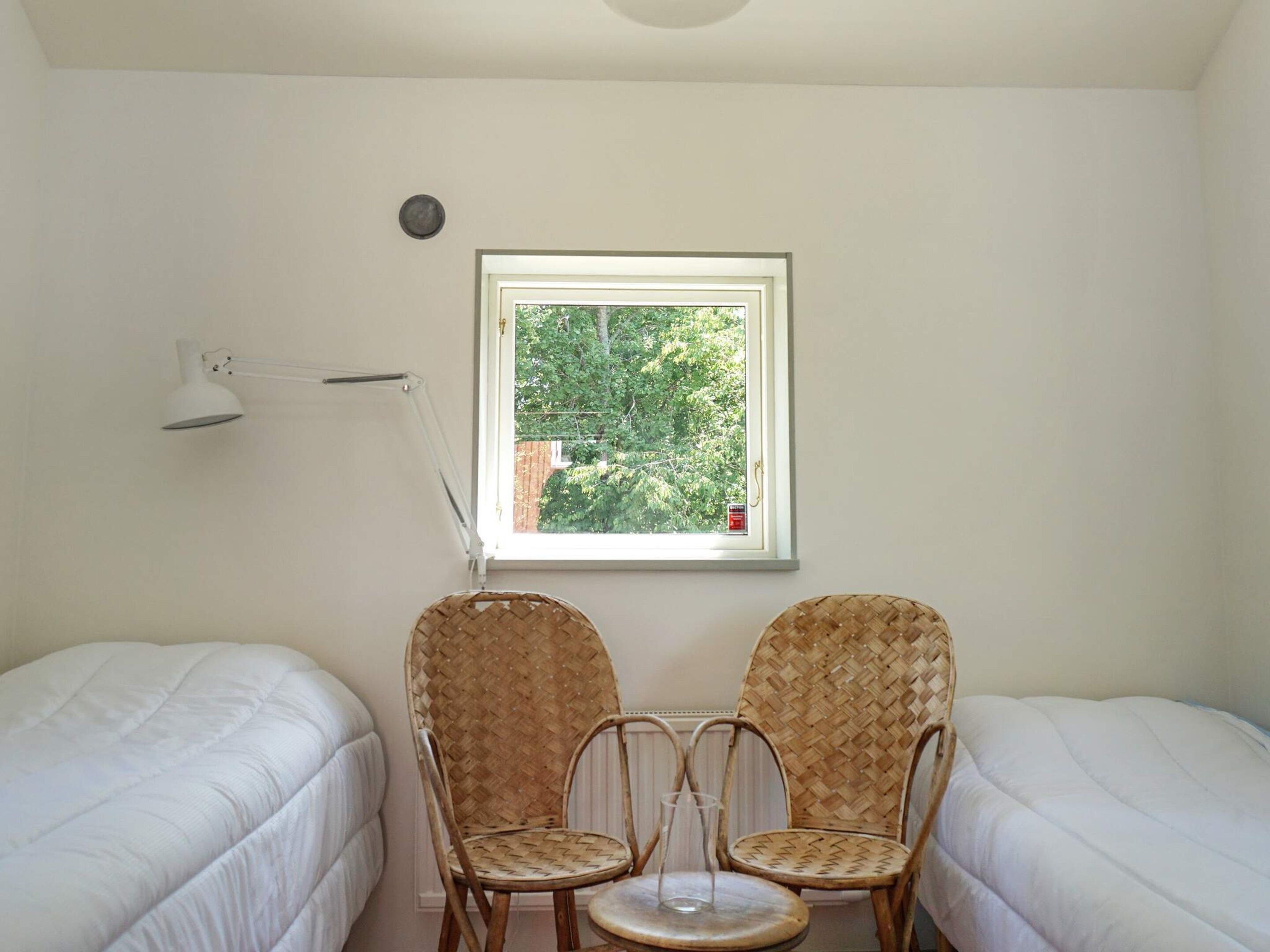 A small, bright room with two single beds, a window with a view of trees, a lamp, and two wicker chairs.