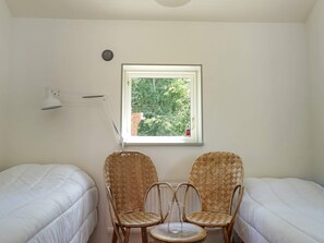 A small, bright room with two single beds, a window with a view of trees, a lamp, and two wicker chairs.
