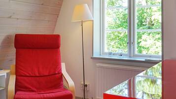 A red chair near a window with a view of greenery, a floor lamp, and a table with a red edge.