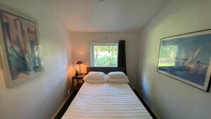 A hotel room with a bed, a window with a view of greenery, a nightstand with a lamp, and a framed picture on the wall.