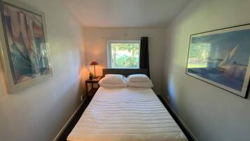 Een hotelkamer met een bed, een raam met uitzicht op groen, een nachtkastje met een lamp en een foto in een lijst aan de muur.