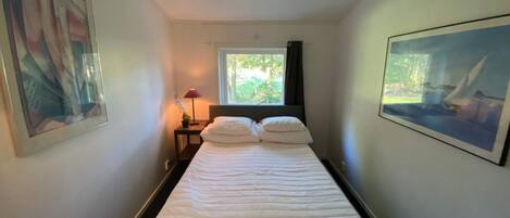 A hotel room with a bed, a window with a view of greenery, a nightstand with a lamp, and a framed picture on the wall.