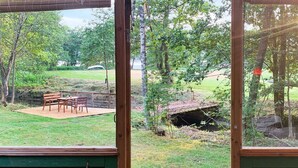 A view through a window of a room with a wooden deck, outdoor seating, and a natural landscape.