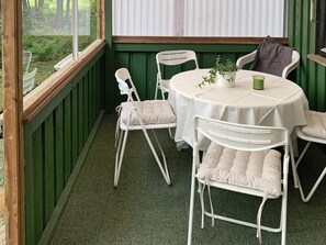 A room with a round table, white chairs, a potted plant, and a view of greenery through a window.