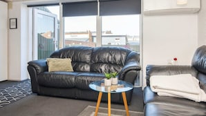 Een moderne woonkamer met een leren bank in zwart, een glazen salontafel en een stadszicht door grote ramen.