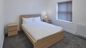 A bedroom with a wooden bed, a bedside table, a nightstand, and a window with blinds. - Apartment 4 in Loftus (Saltburn-by-the-Sea)