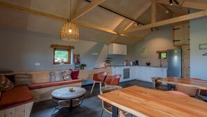 A cozy, rustic interior with a wooden ceiling, a dining area with a round table and chairs, a built-in bench with cushions, and a kitchen area with cabinets and appliances. - Lodge in Drenthe With Scenic Veranda (Ruinerwold)