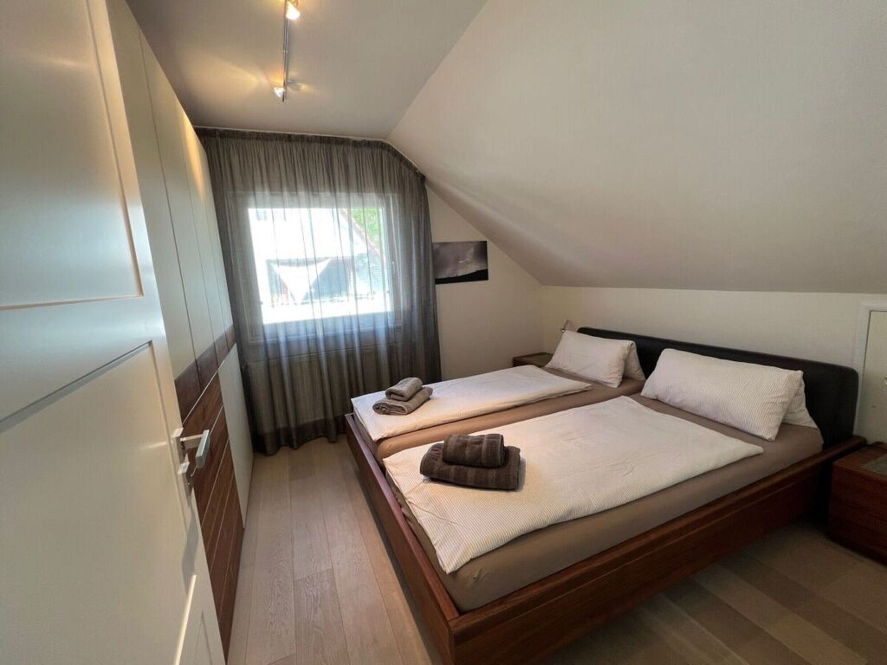 A bedroom with two beds, a window with curtains, a bedside table, and a small painting on the wall.