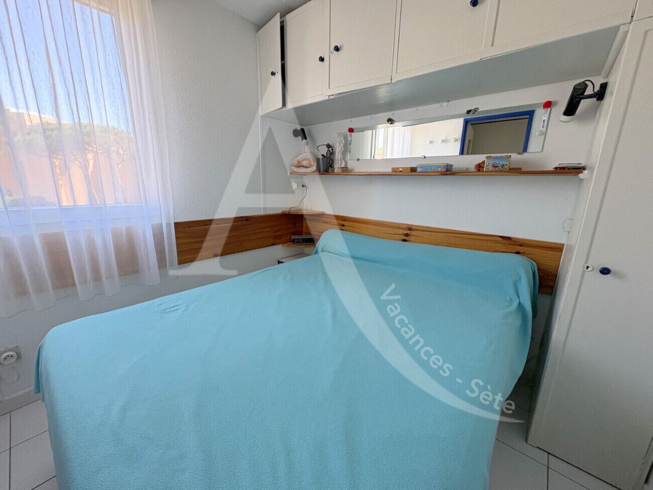 A bedroom with a bed, a wooden headboard, a window with blinds, a white wardrobe, and a shelf with various items.