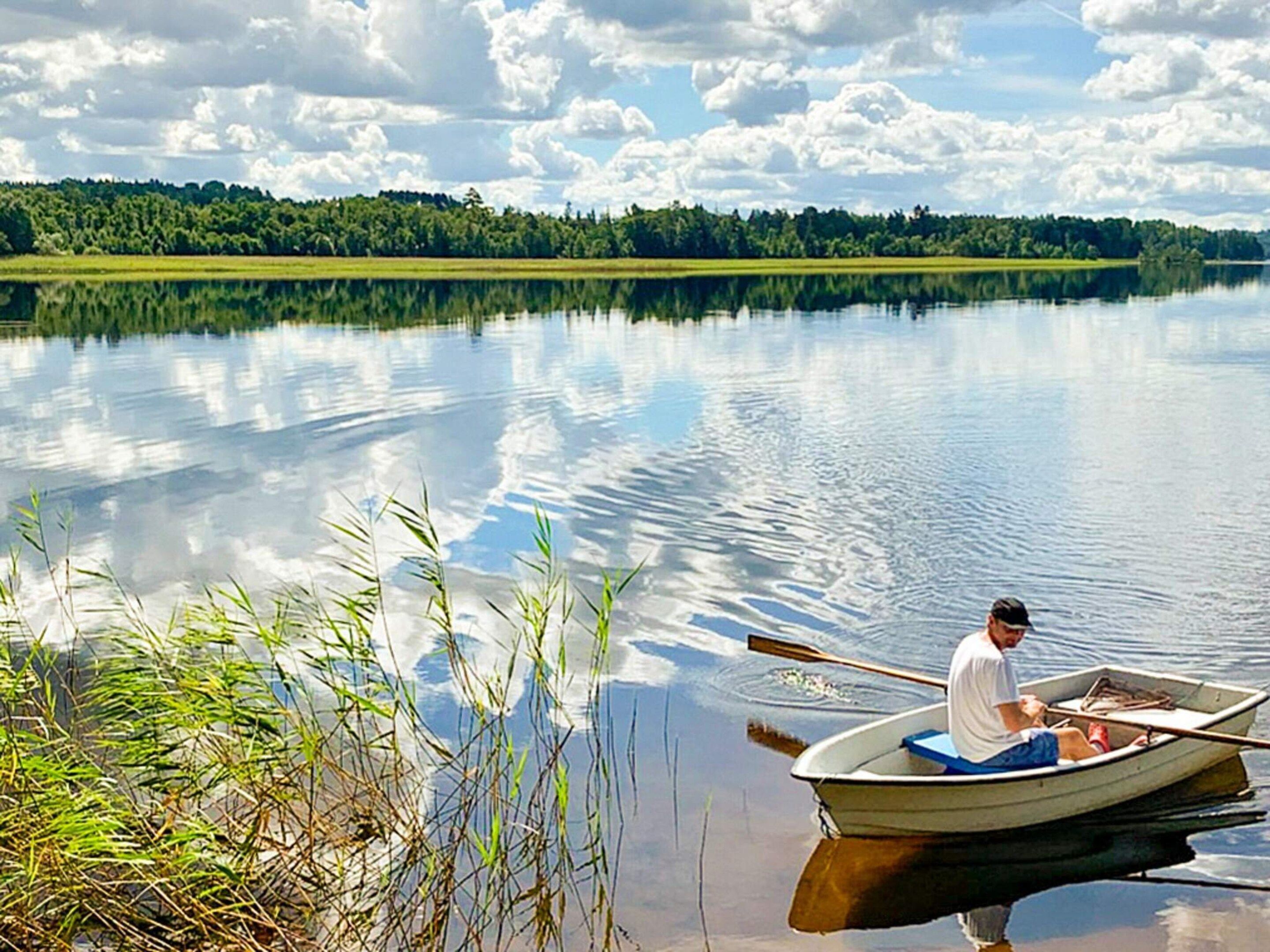 Un lac paisible, avec une seule barque à rames et une personne qui rame, le tout entouré de verdure et d’arbres.