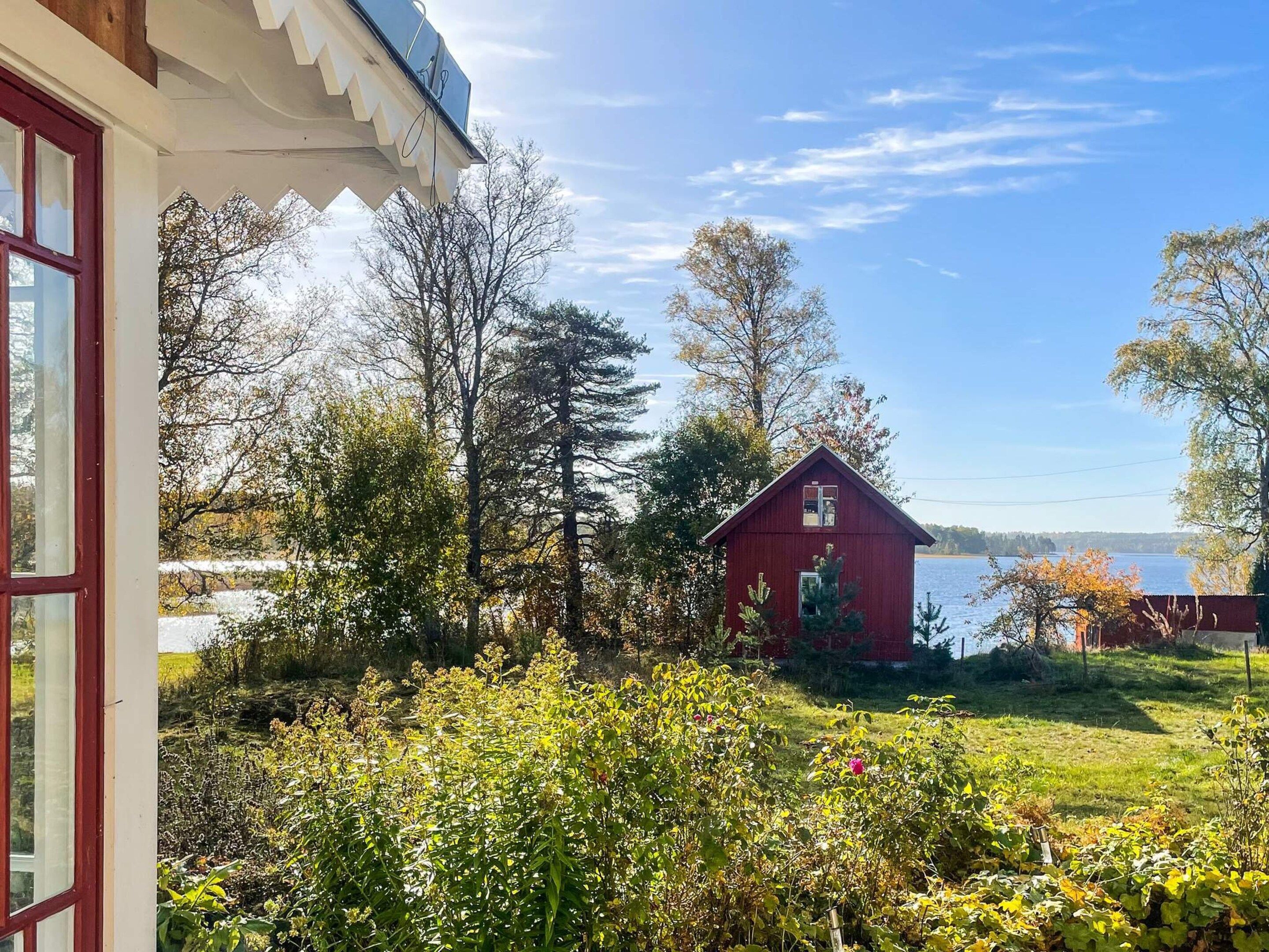 A red wooden house with a gabled roof, surrounded by lush greenery and trees, with a body of water in the background.