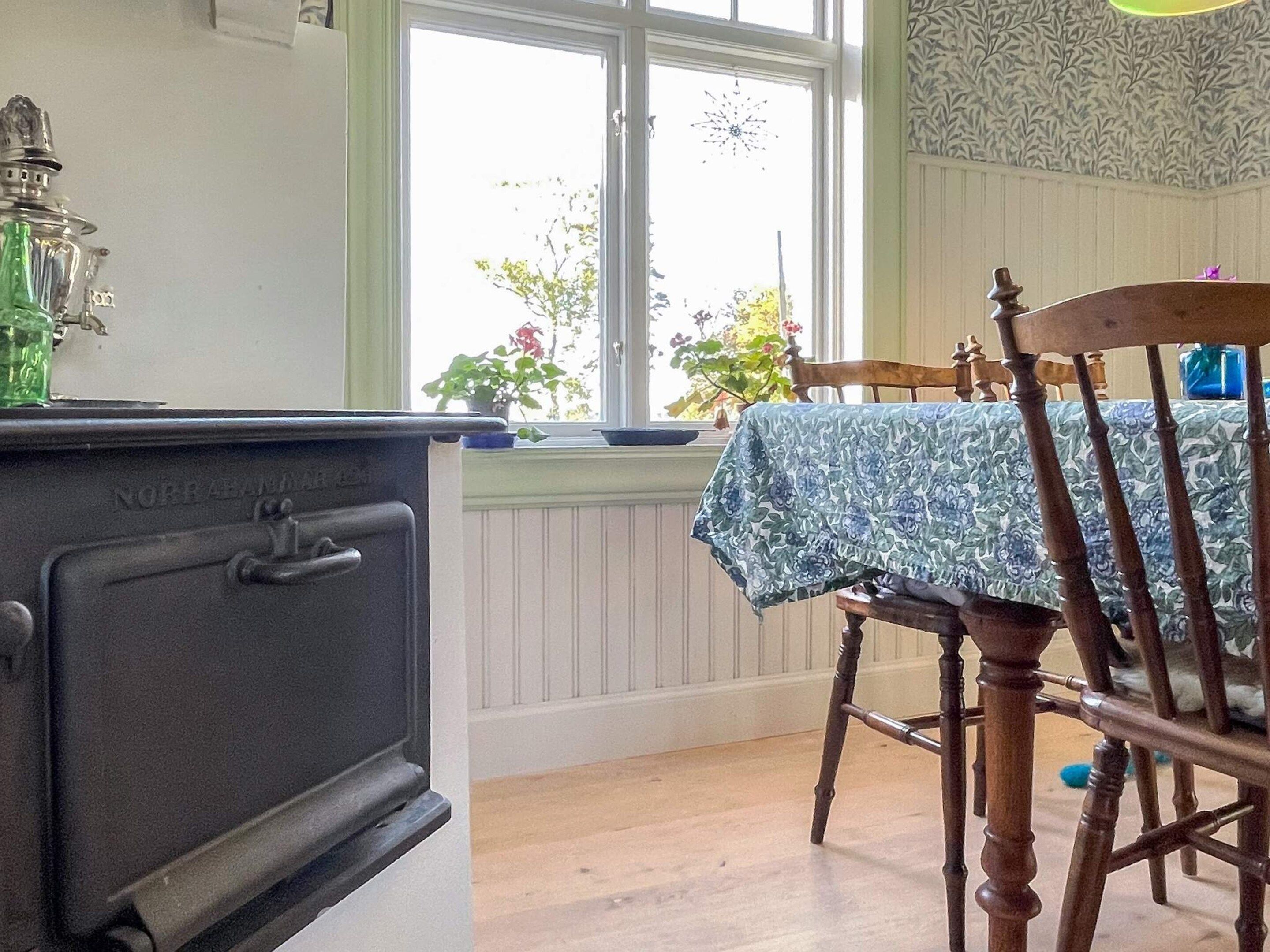 Eine Küche mit einem alten Herd, einem Fenster mit Blick auf Grünflächen und einem Tisch mit einer Tischdecke mit Blumenmuster.