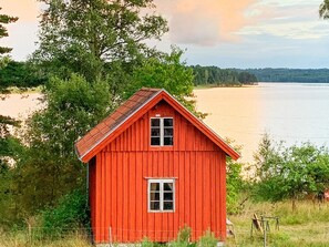 Een rode houten hut met een puntig dak, omringd door bomen en een waterpartij.