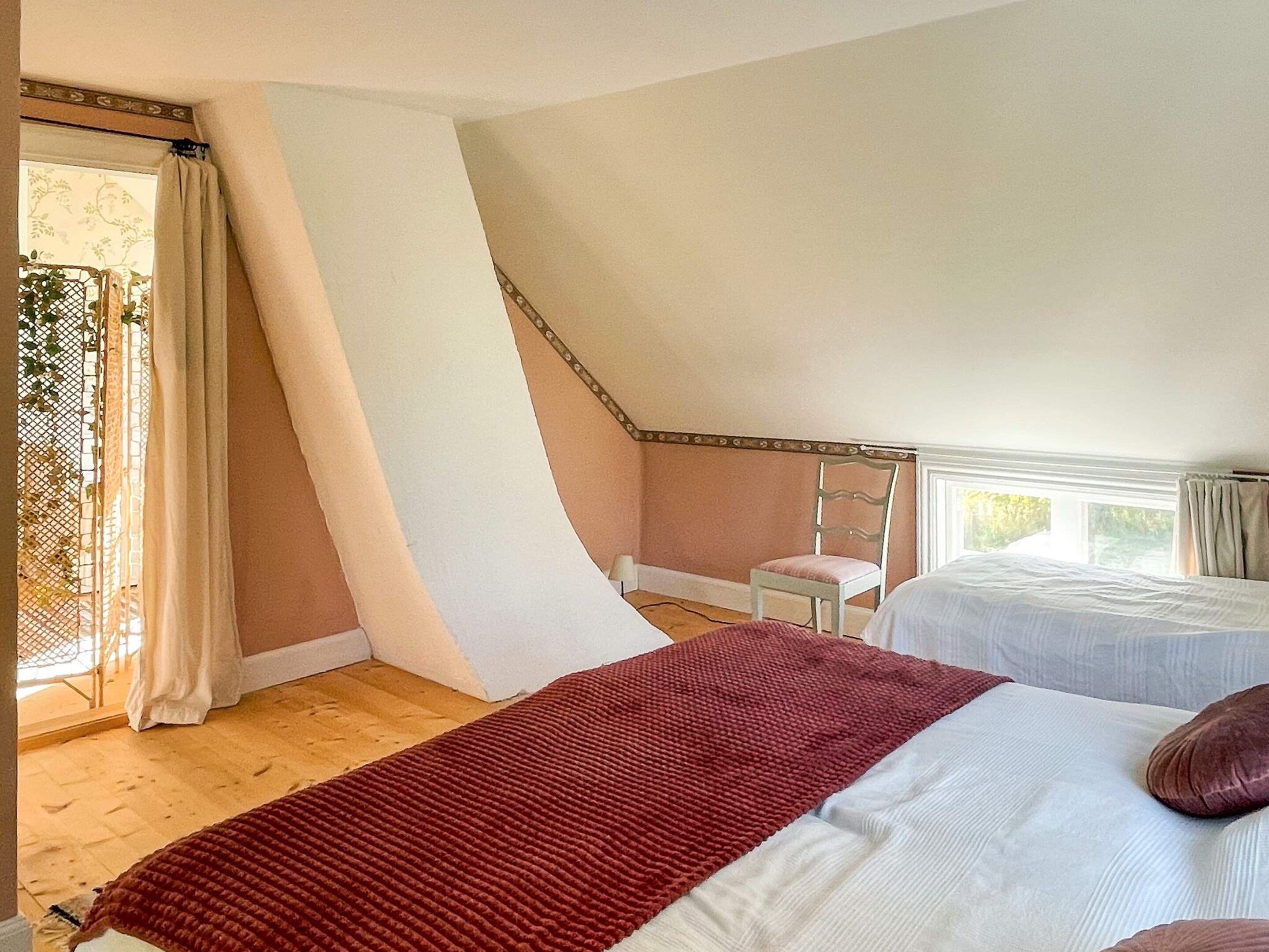 A bedroom with two beds, a wooden floor, a window with curtains, and a chair.