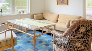 Een kamer met een bank, een tafel met glazen blad, een rieten stoel en een raam met uitzicht op bomen.