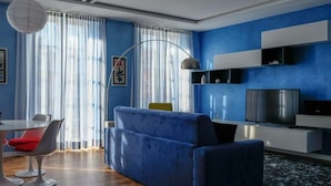 Uma sala de estar moderna com um sofá azul, uma mesa de jantar com cadeiras, uma televisão e uma grande janela com cortinas.