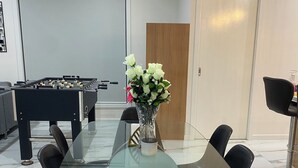 Un espace repas moderne avec une table en verre entourée de chaises noires aux pieds en bois, un vase avec des fleurs blanches et, au fond, une table de baby-foot.