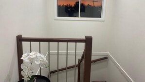 Un escalier avec une rampe en bois, un pot d’orchidée blanche et une fenêtre donnant sur un coucher de soleil et des arbres se découpant en silhouette.