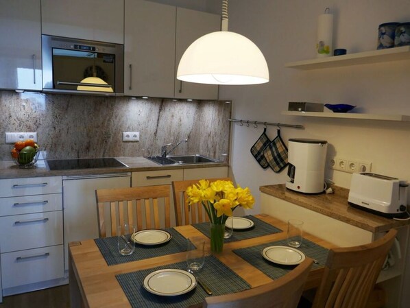 A compact kitchen with a built-in oven, a sink, and a toaster. There is a dining table with chairs and a vase of yellow flowers. - Ostseeblick Berolina (Dahme)