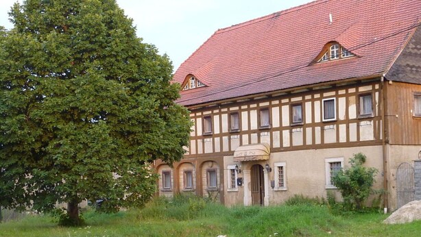 A traditional half-timbered building with a red-tiled roof, a large tree in front, and a stone wall to the left. - Saalendorf - Holidays (Grossschoenau)