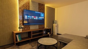 A modern living room with a flat-screen TV mounted on a textured wall, a wooden entertainment unit, a round coffee table, and a beige sofa. - XcellHIROSHIMA HEIWAODORI (Hiroshima)