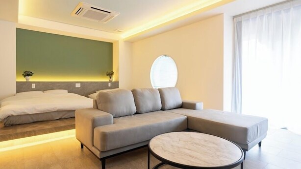 A modern hotel room with a sectional sofa, a round coffee table, and a bed with a green accent wall. - XcellHIROSHIMA HEIWAODORI (Hiroshima)