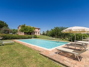 Una piscina rectangular con solárium empedrado, rodeada de césped y un muro de piedra. Se observan sillas de descanso y un gran parasol.