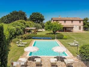 Piscina rectangular con dos sombrillas, rodeada por una terraza de piedra y sillas de descanso.
