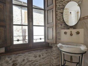 A bathroom with a stone floor, a wooden door, a window with a view of a building outside, a round mirror, and a sink with a stone countertop. - Terrinca Suite Comfortable Holiday Residence (Stazzema)