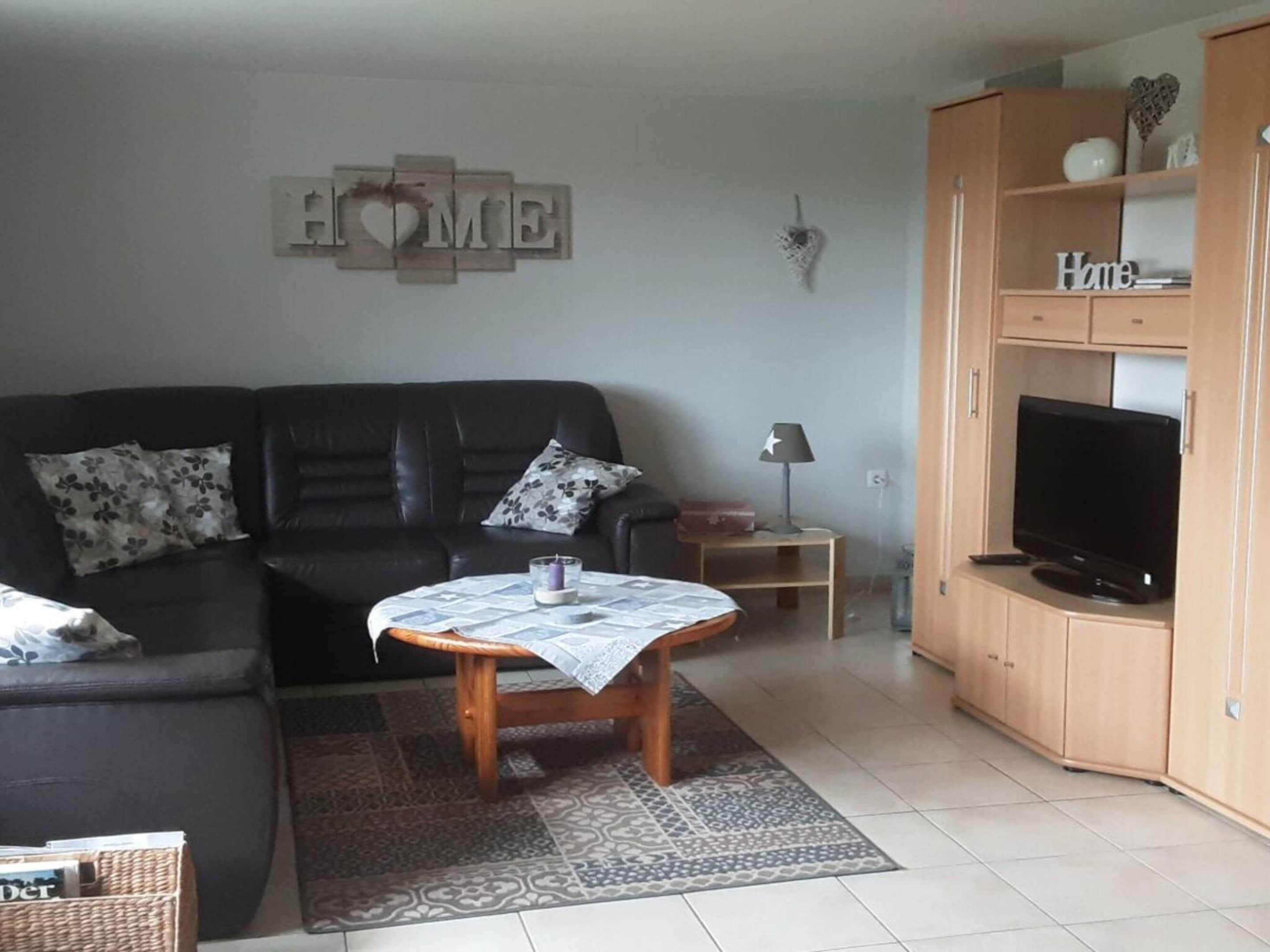 A living room with a black leather sofa, a wooden coffee table, a television on a wooden cabinet, and a 'HOME' sign on the wall.