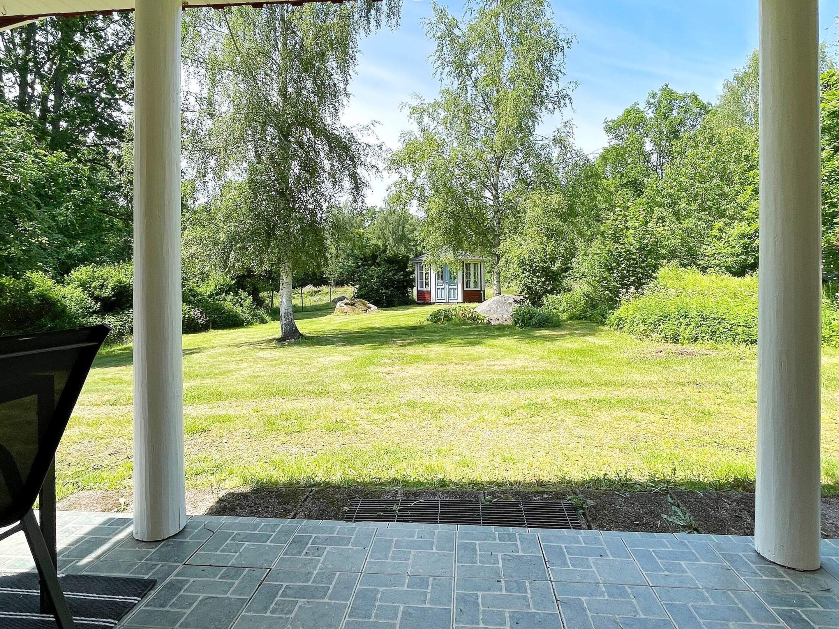 En täckt uteplats med utsikt över en gräsyta, träd och en liten byggnad.