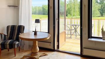A room with a wooden floor, a striped chair, a round table with a lamp, and a view of a balcony with outdoor furniture.