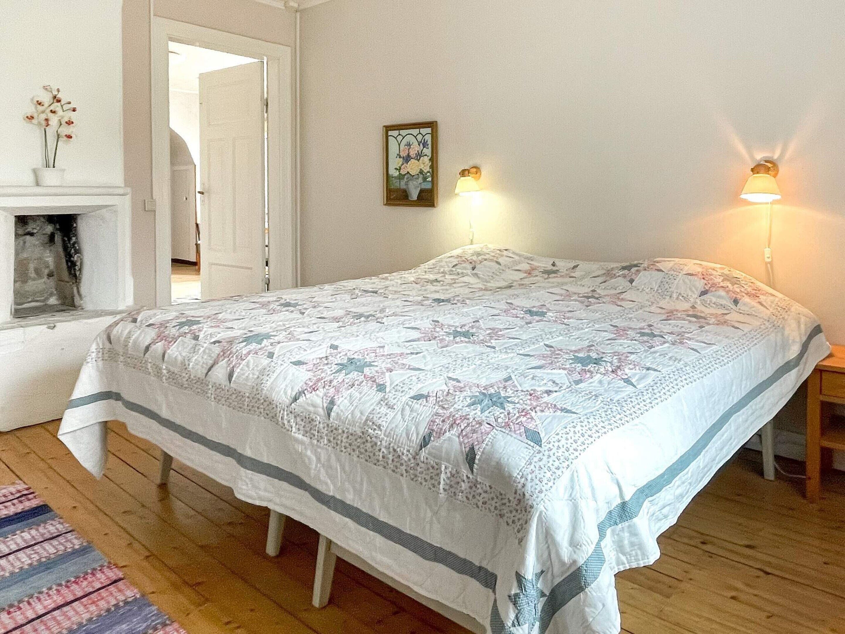 A bedroom with a large bed, a fireplace, two wall-mounted lamps, and a door leading to another room.